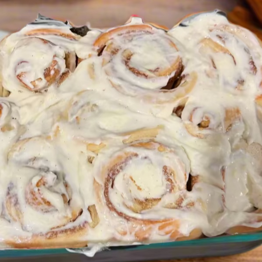 Glass dish with cinnamon rolls covered in white frosting on a marble surface with cinnamon sticks.